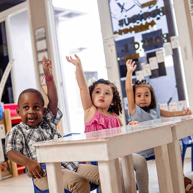 Three young children sitting at a desk in a classroom all raising their hands